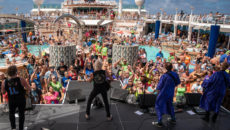 80's band on an outside stage on a cruise ship with a large crowd and a pool in the background.